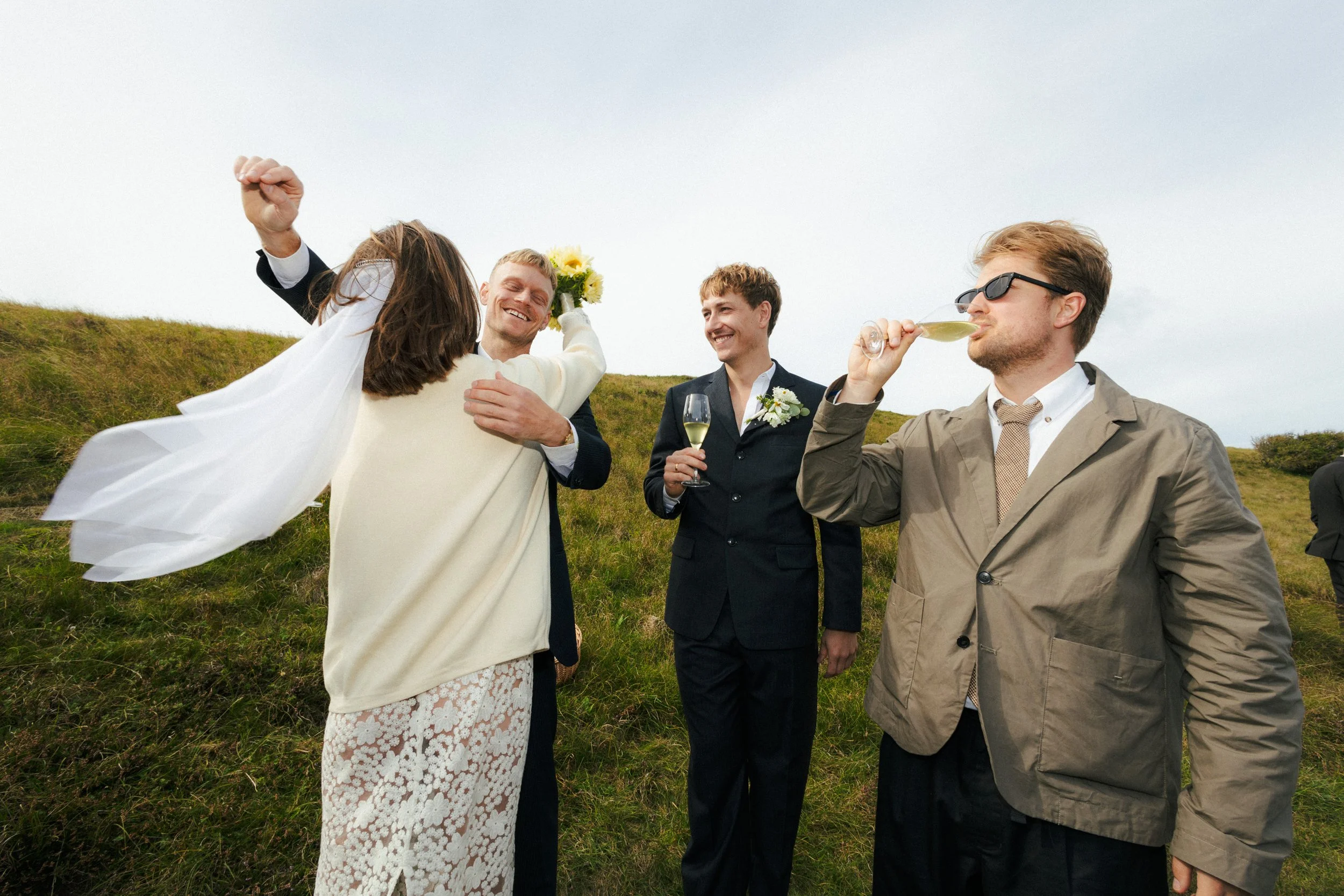 Gruppe af mænd og kvinder i formelle tøj, der fejrer ude i det grønne med skum, blomster og champagne.