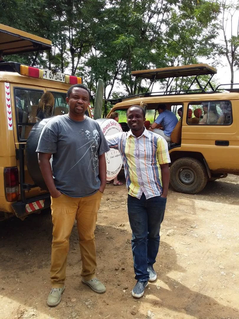 Two men standing in front of safari vehicles with other people inside, surrounded by trees.