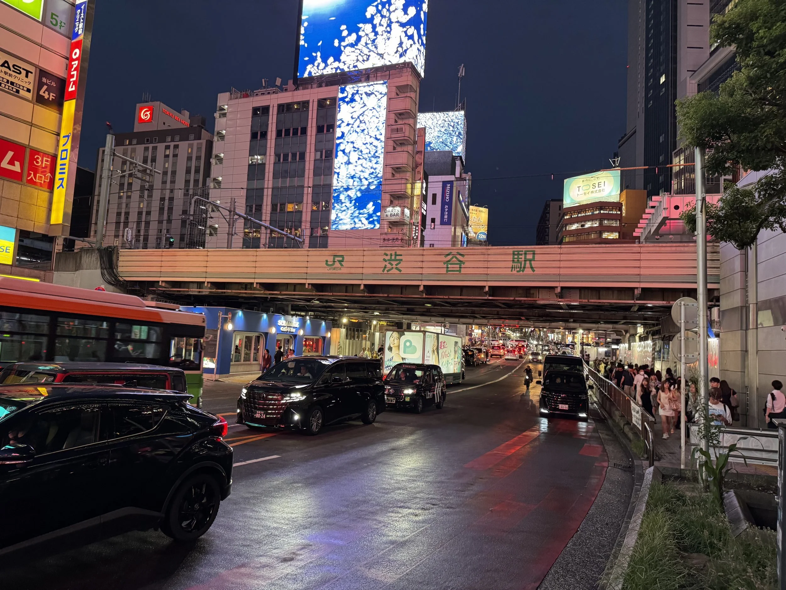 A street at night by a railway bridge in Tokyo, lit by large digital screens on the buildings above, with taxis on a wet road and people walking on the pavement.