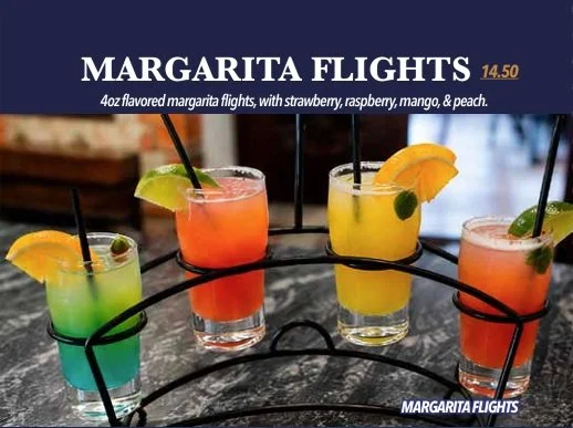 Cuatro vasos de margaritas de sabores diversos sobre una mesa de piedra, con fruta y pajillas negras, en un soporte circular metálico.