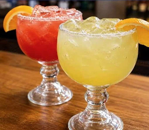 Dos cócteles en copas cortas con hielo y rodajas de naranja, uno de color rosa y otro amarillo, sobre una mesa de madera.