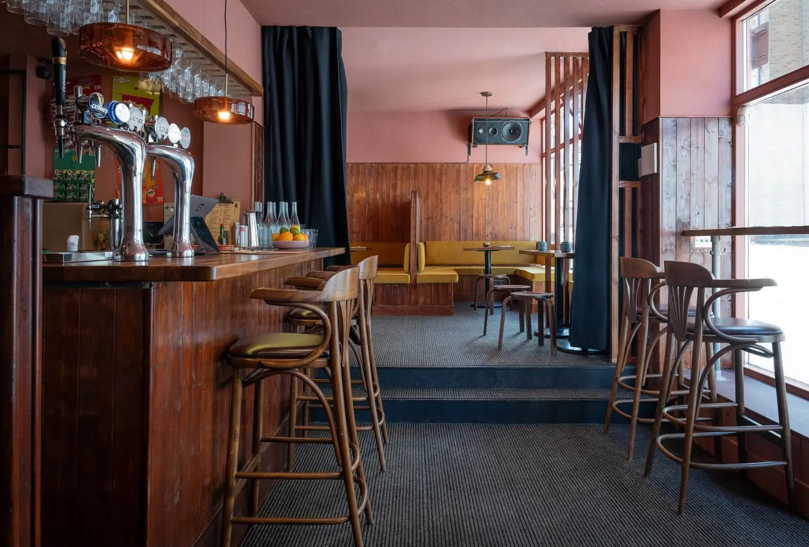 Interior of a cozy cafe with wooden furniture, a bar counter with taps, stools, and yellow seating area near windows with black curtains.