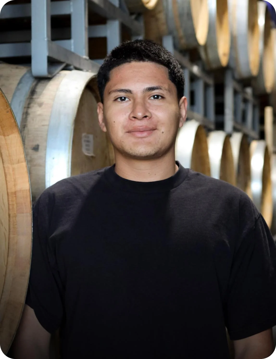 A young man with short black hair and a black T-shirt standing in a wine cellar, surrounded by large wooden barrels.