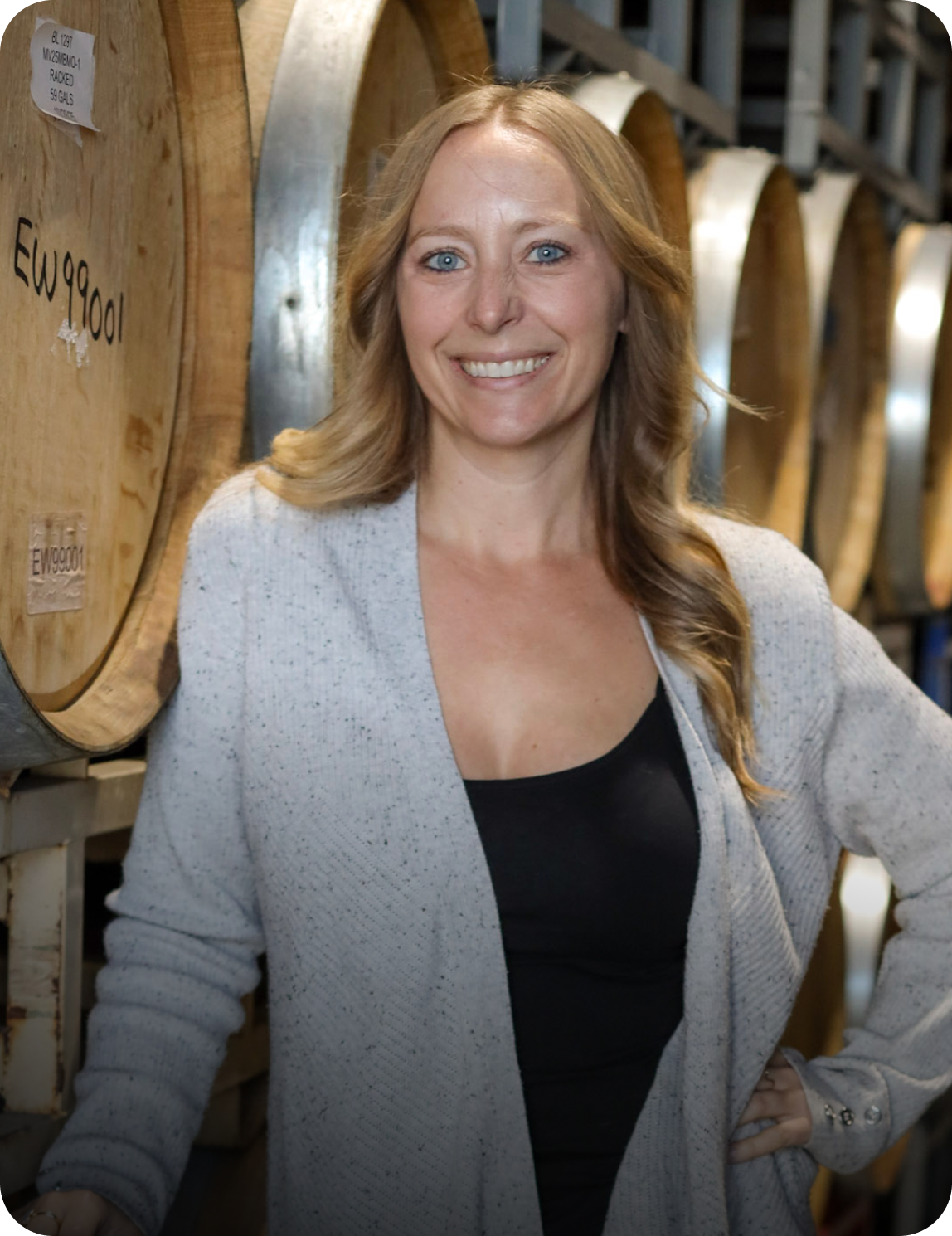 A woman with long wavy blonde hair and blue eyes smiling at the camera, standing in a storage room with large wooden wine barrels behind her.
