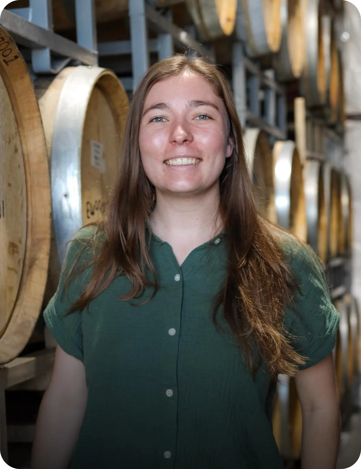 A young woman with long brown hair smiling in a cellar among wooden wine barrels.