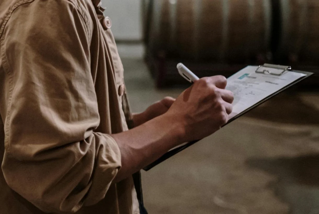 Person in beige jacket writing on a clipboard with a white pen.