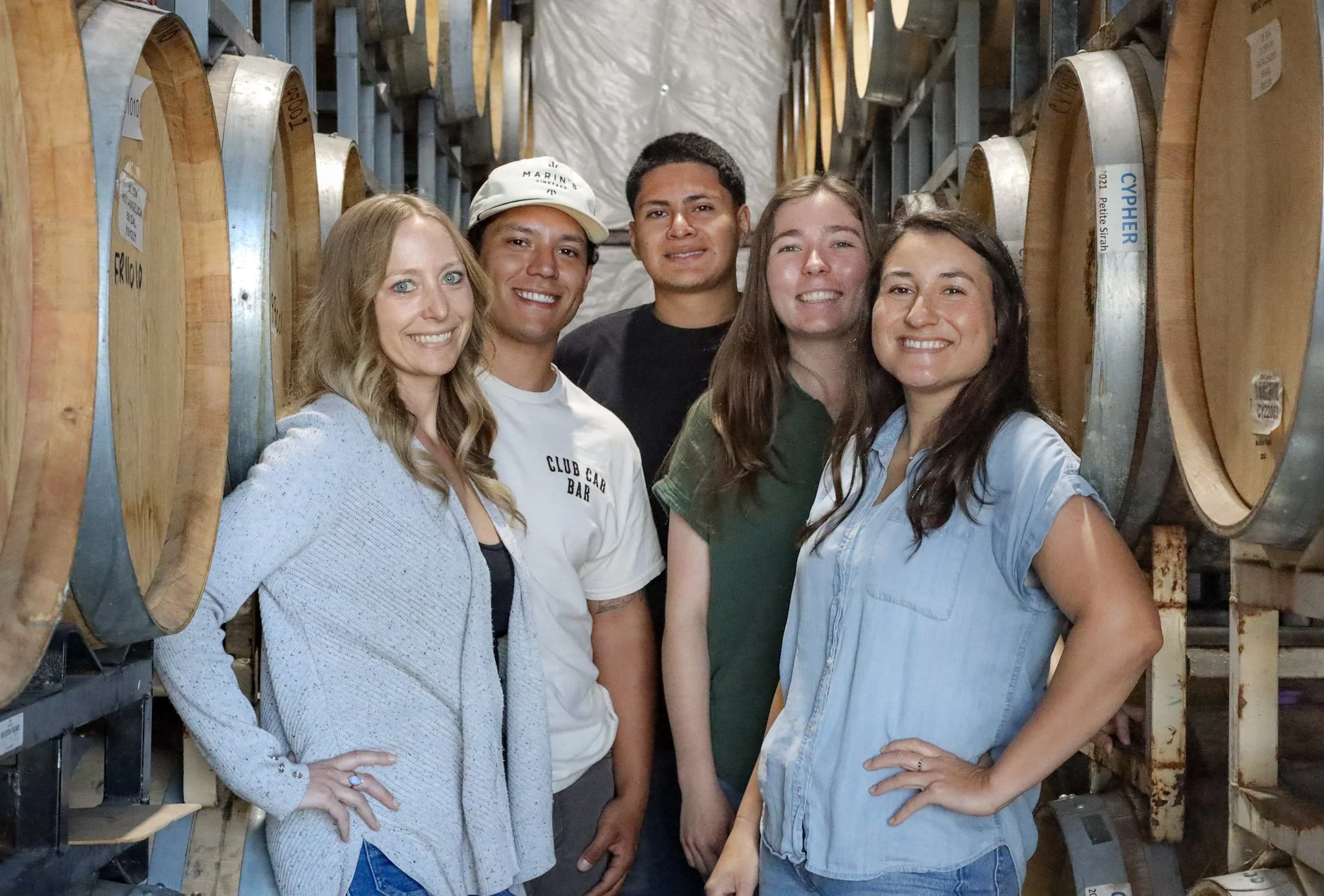 A group of five smiling people standing close together inside a warehouse or storage room with large wine barrels on shelves around them.