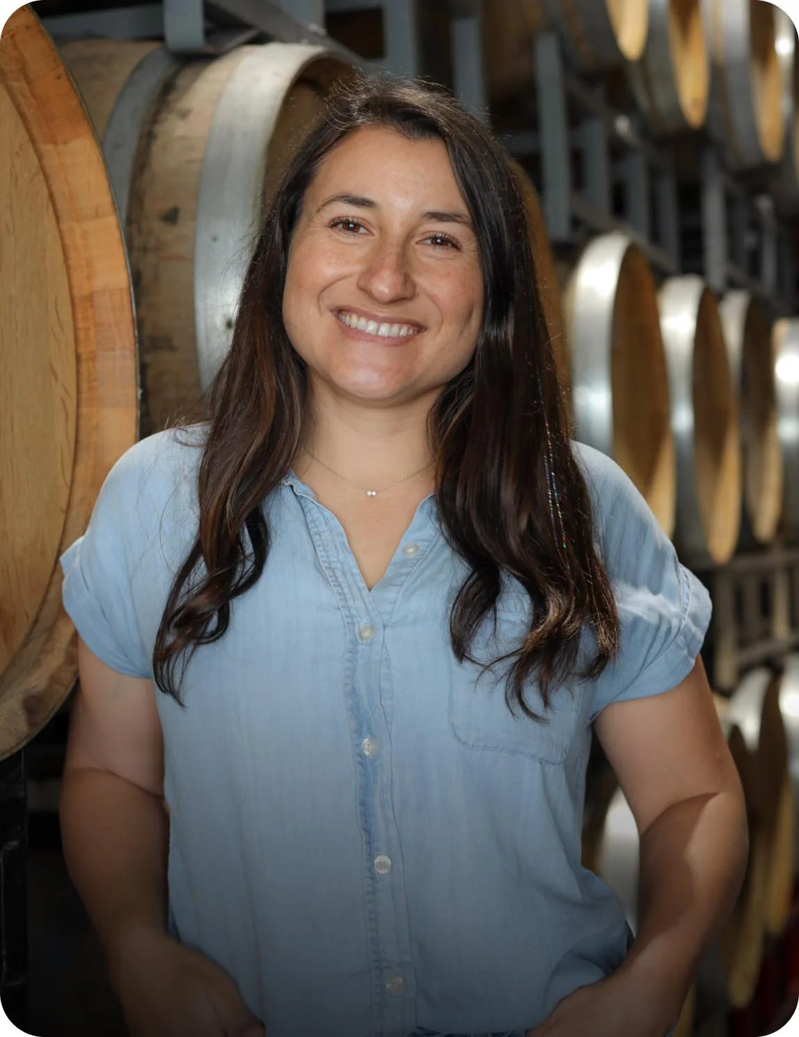 A woman with long dark hair smiling, wearing a light blue shirt, standing in front of wine barrels in a winery or wine cellar.