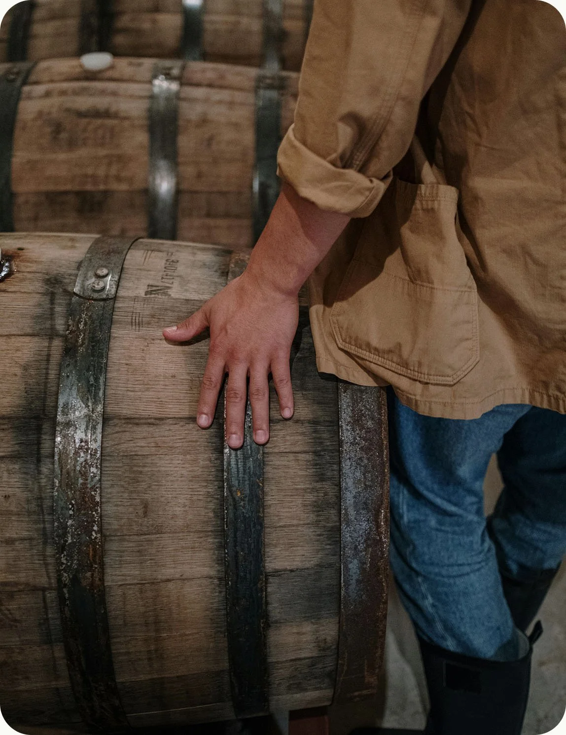 A person in a tan jacket and jeans touching a wooden wine barrel with a metal hoop.