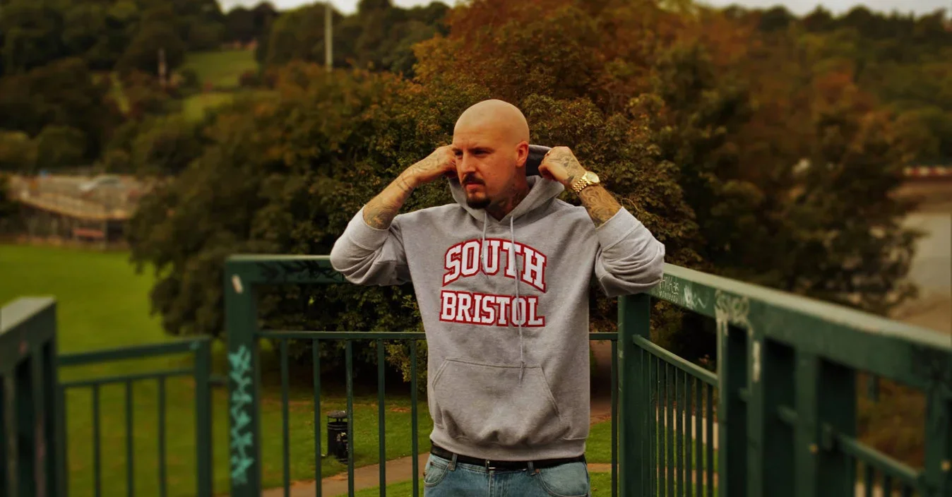 Bald man with tattoos adjusting hoodie on a green bridge overlooking a park with trees and grass.
