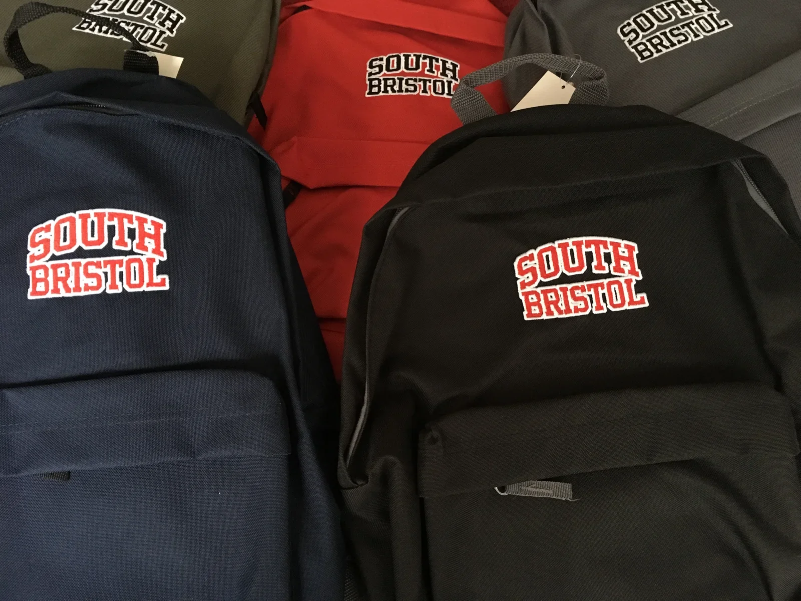 Four backpacks with 'South Bristol' embroidered on them, two are black, one is navy blue, and one is red, all on a surface.