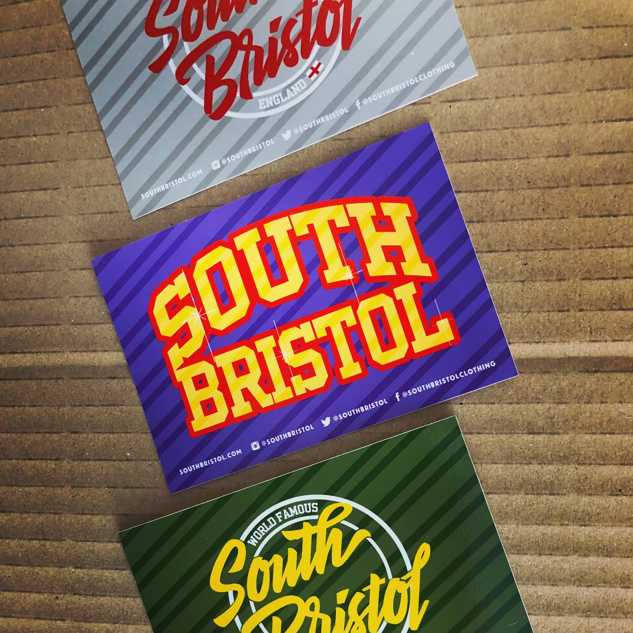 Close-up of colorful promotional posters on a wooden surface. The top poster features red and white text with social media icons and a website address on a gray and white striped background. The bottom poster has yellow and red text on a purple background with striped pattern.