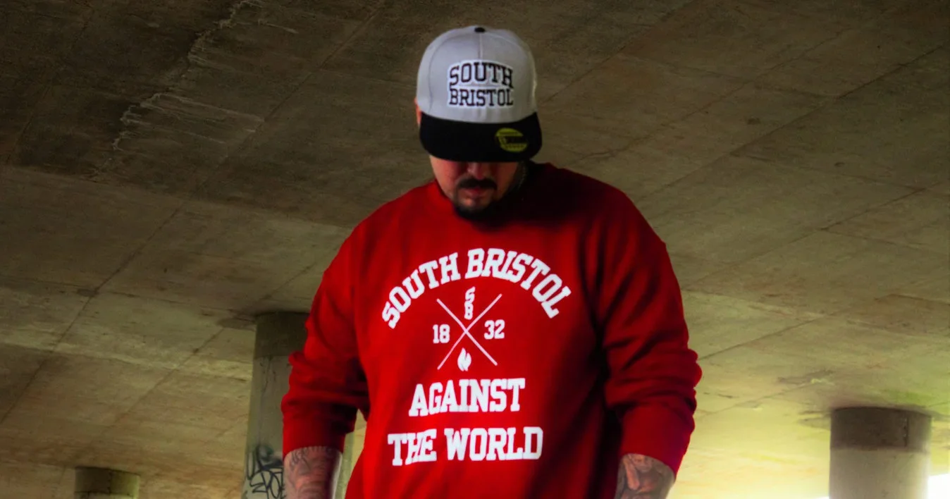Man wearing a red sweatshirt with white text, a black and white baseball cap, and tattoos on his arms under a concrete bridge.