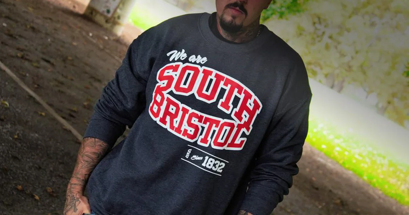 Person wearing a gray sweatshirt with red and white text that says "We are SOUTH BRISTOL" and "Est. Since 1832" standing outdoors on a dirt path with trees in the background.