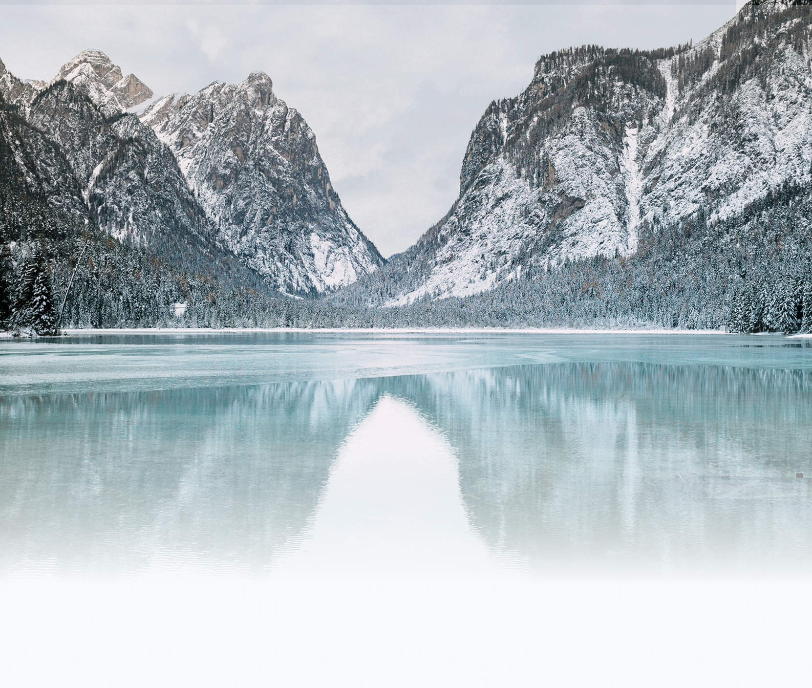 Snow-covered mountains surrounding a partially frozen lake with a reflection of the mountains and cloudy sky.