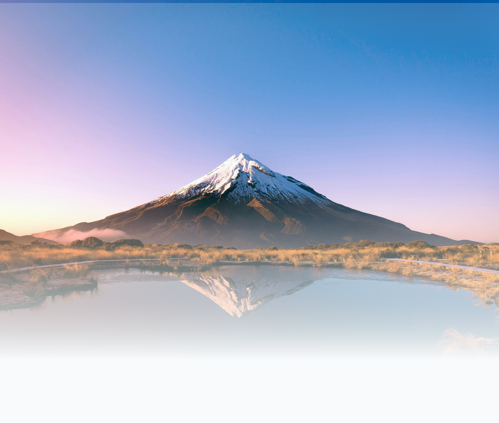 Snow-capped mountain reflected in a calm body of water with a clear blue sky and a grassy area in the foreground.