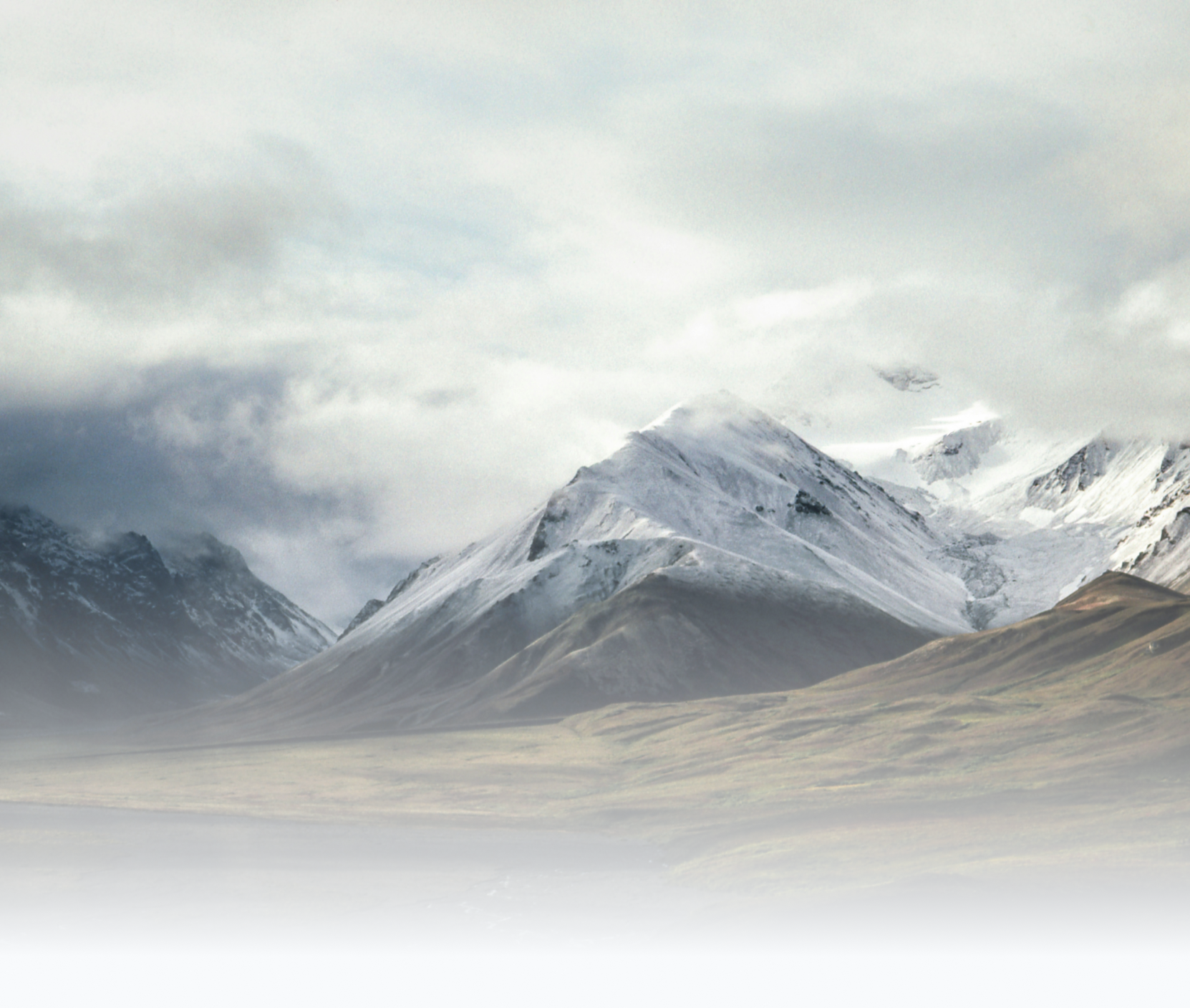 Snow-covered mountains in a cloudy sky with grassy plains in foreground.