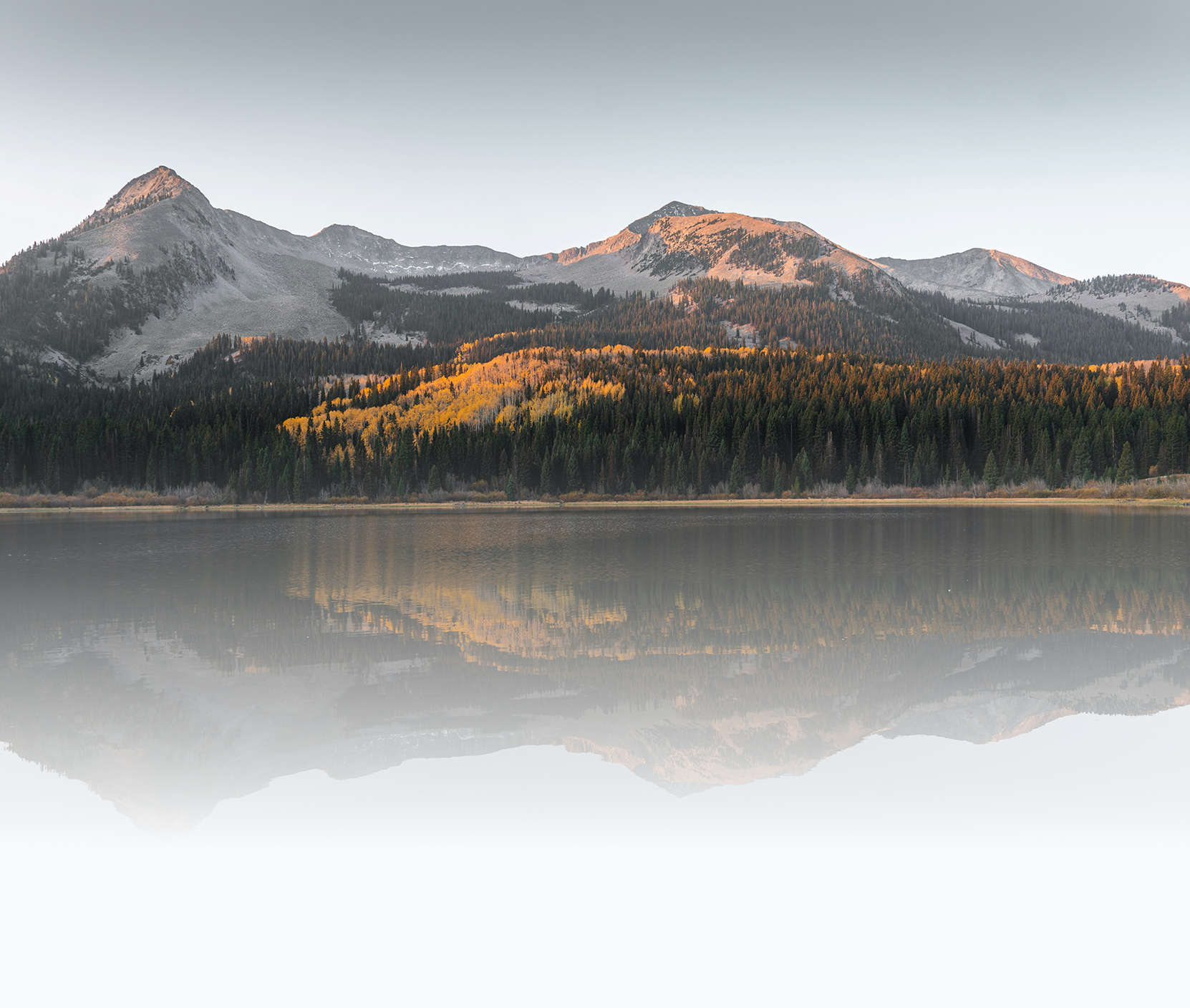 Scenic view of a mountain range reflected in a calm lake, with forested slopes and a clear sky.