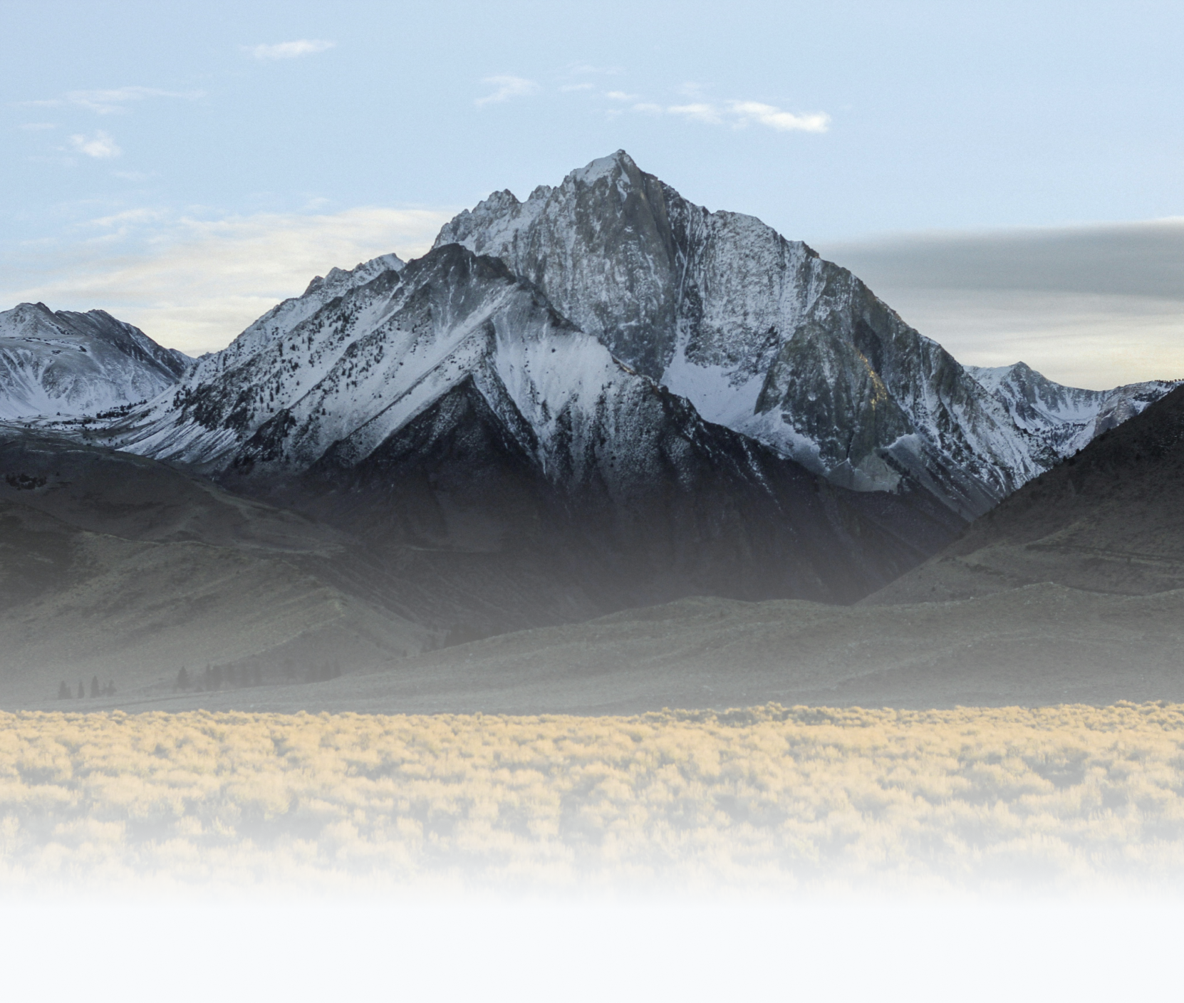 Snow-capped mountain with rugged peaks rising above a haze-covered valley under a partly cloudy sky.