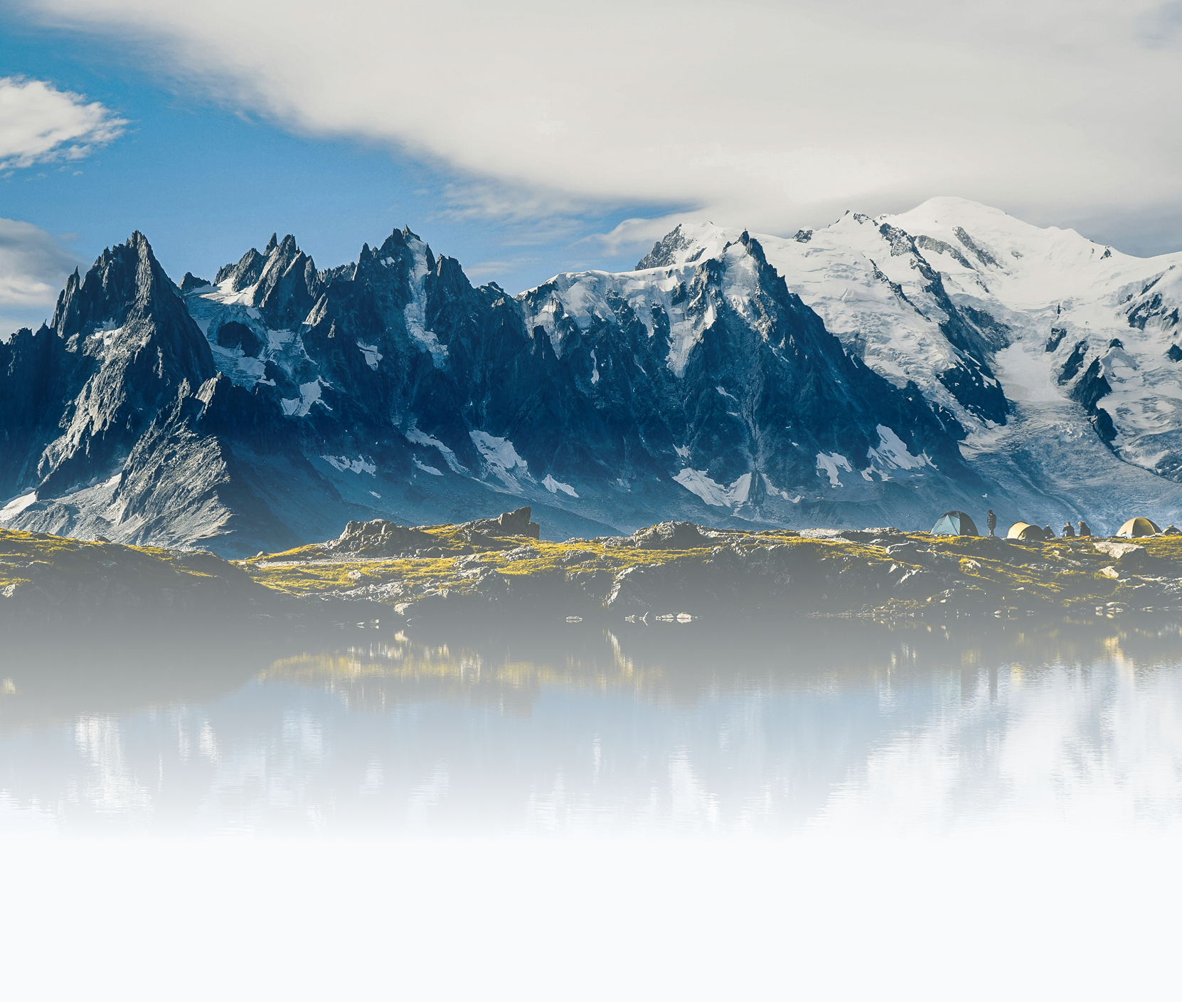 Snow-capped mountains with jagged peaks and a glacier, a group of tents with people nearby, and a calm reflective body of water in the foreground under a partly cloudy sky.