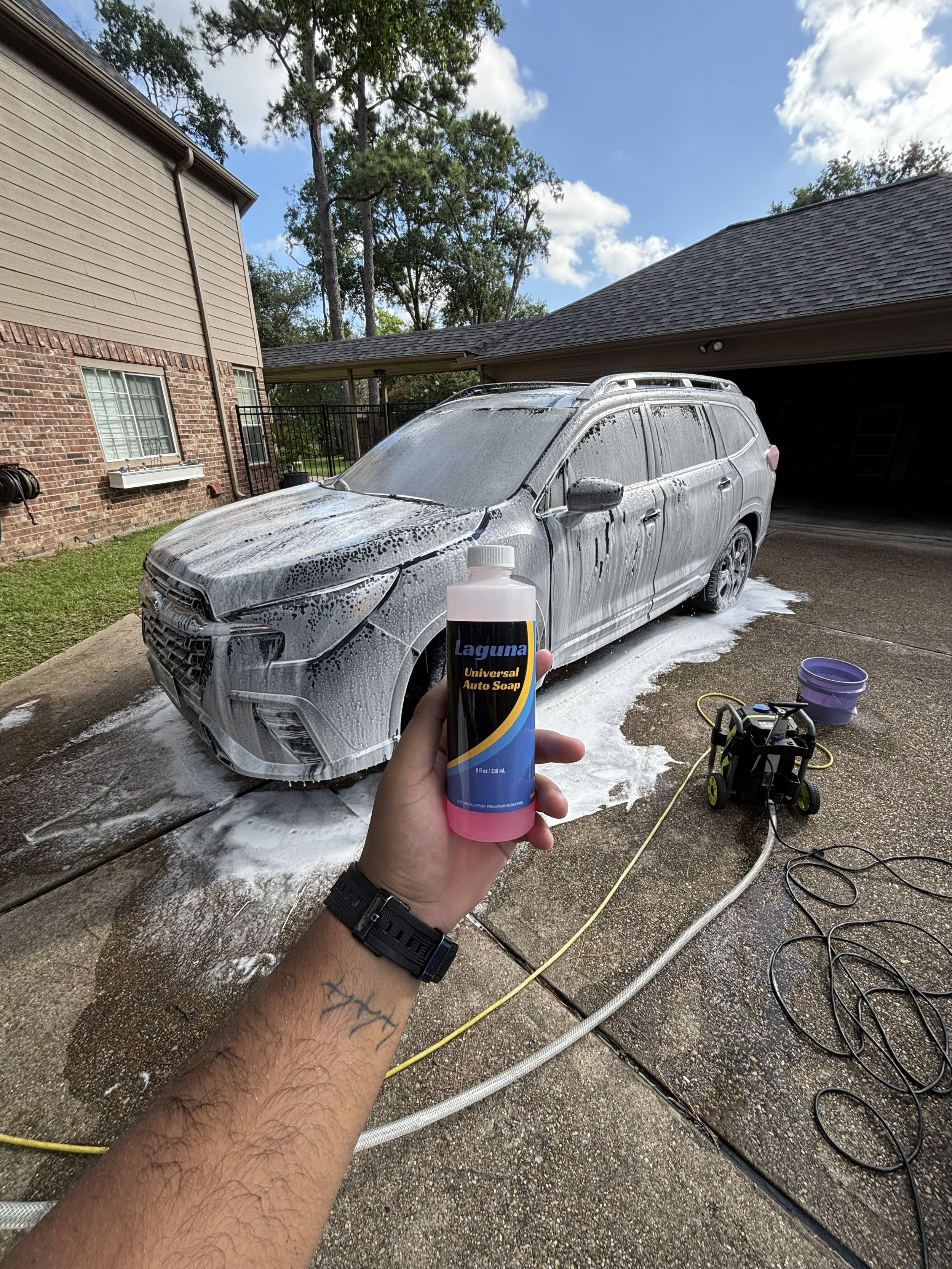 A person holding a bottle of Laguna Universal Auto Soap in front of a car that is covered in soap and being washed with water and foam. The scene is outdoors in a garage driveway with a brick house, a carport, and trees in the background.
