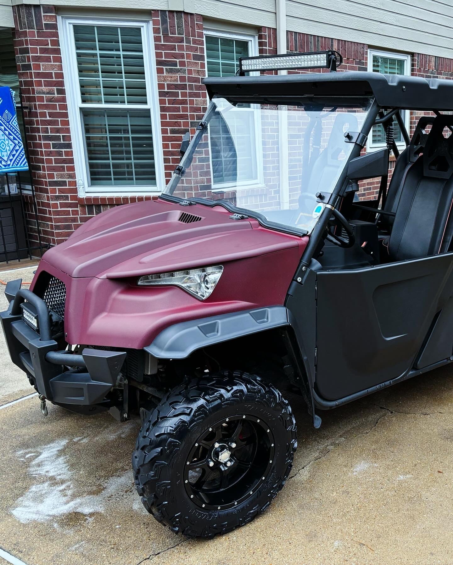 A red and black utility off-road vehicle parked on a wet concrete driveway in front of a brick house with white window frames.