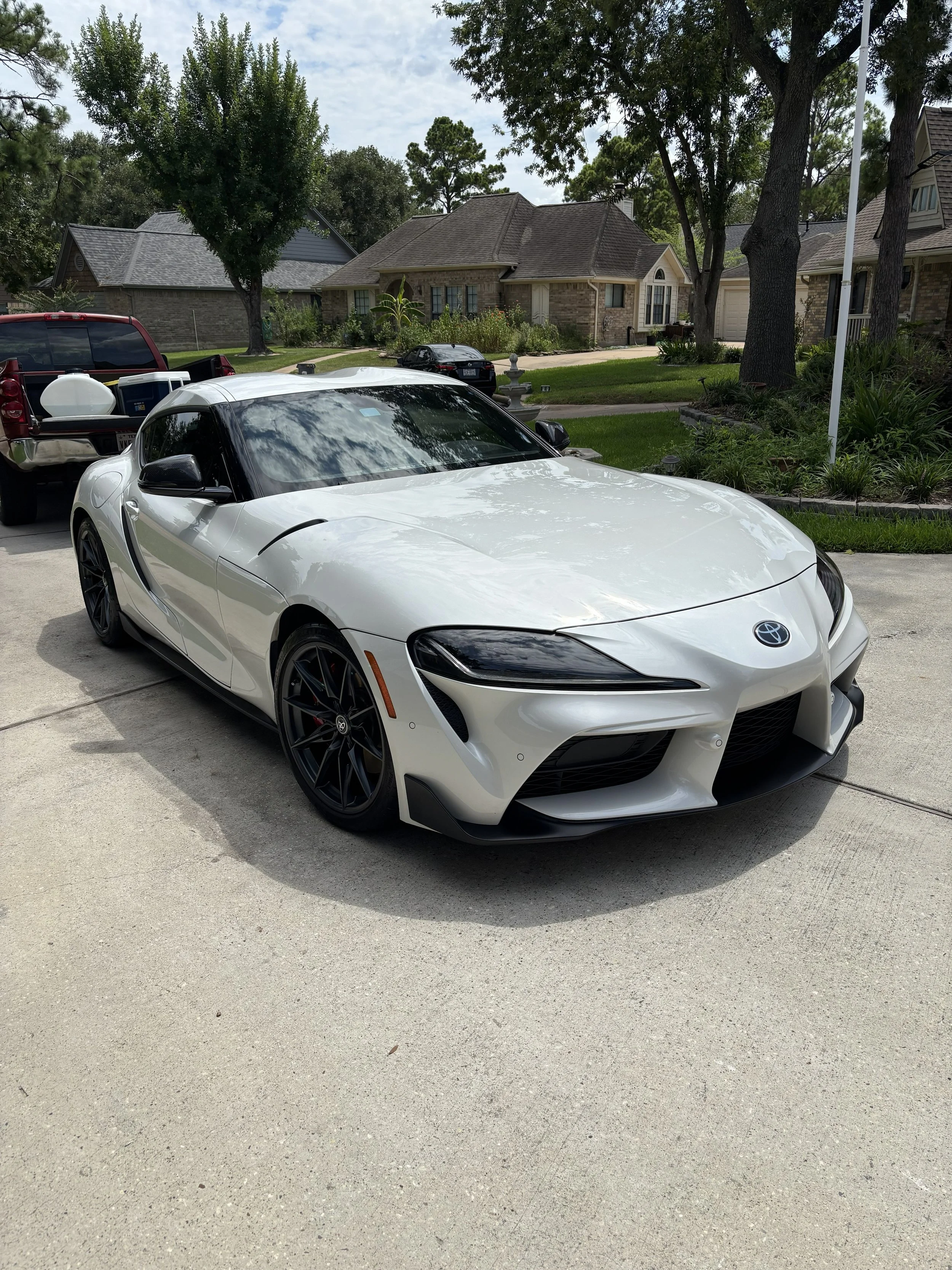 A white sports car parked in a residential driveway with houses and trees in the background.