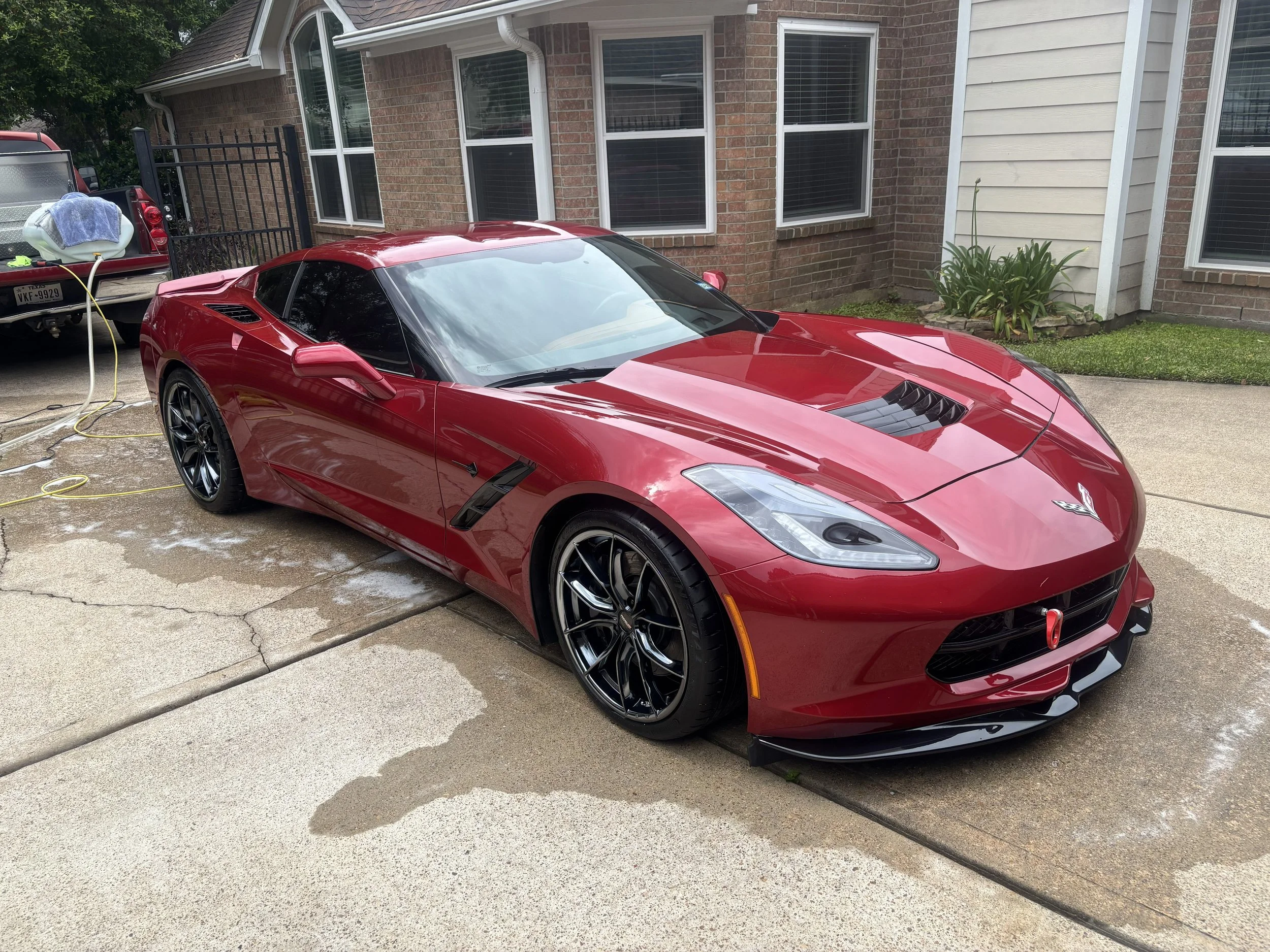 Red sports car parked in driveway near a house with brick and siding exterior, and large windows. Car is being washed with water and soap, with cleaning tools visible nearby.