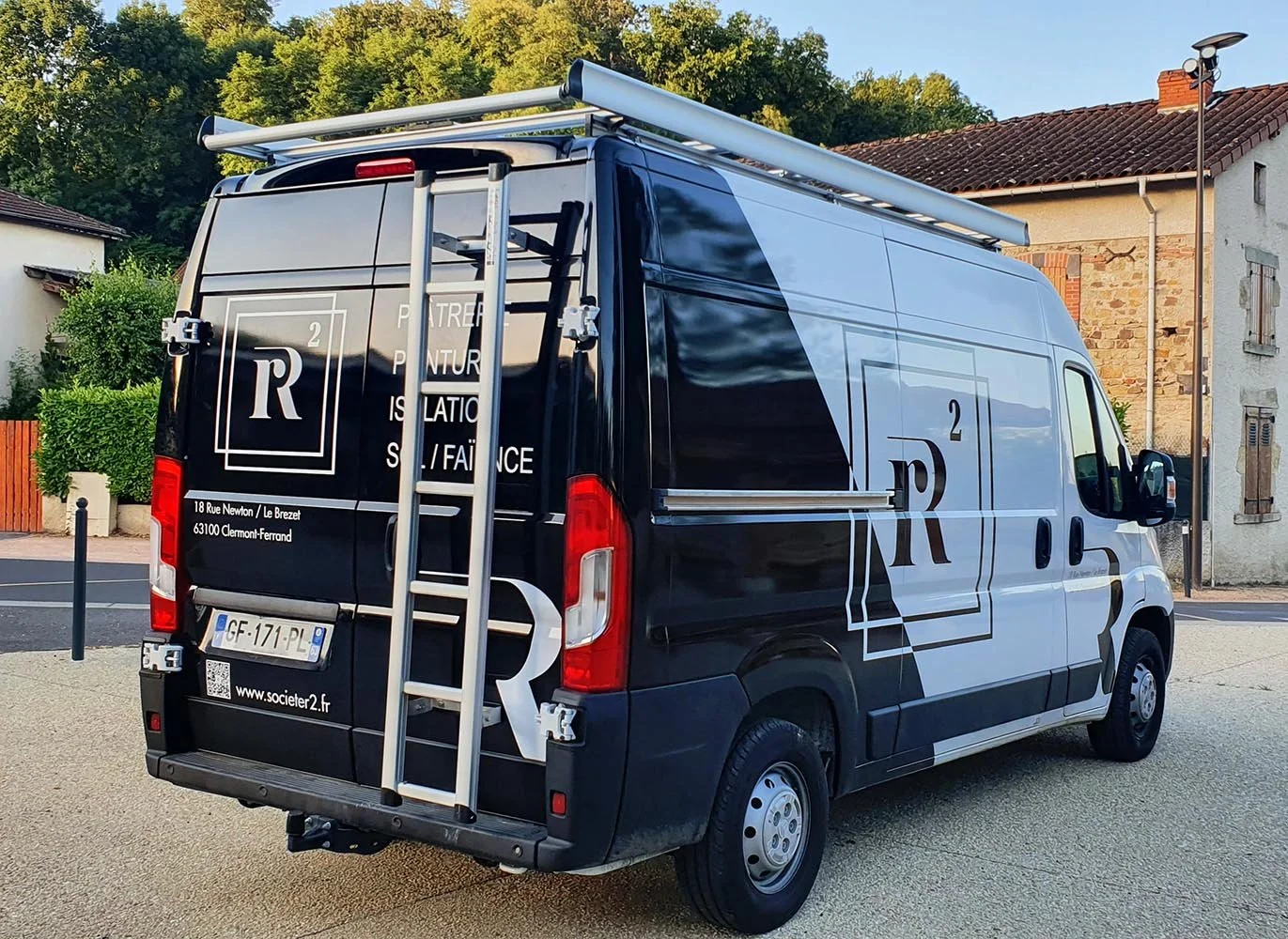 Une camionnette noire avec des logos et textes en blanc, stationnée dans une zone résidentielle avec des maisons en arrière-plan. La camionnette possède une échelle pliante attachée à l'arrière et une rampe de toit, et affiche une plaque d'immatricul