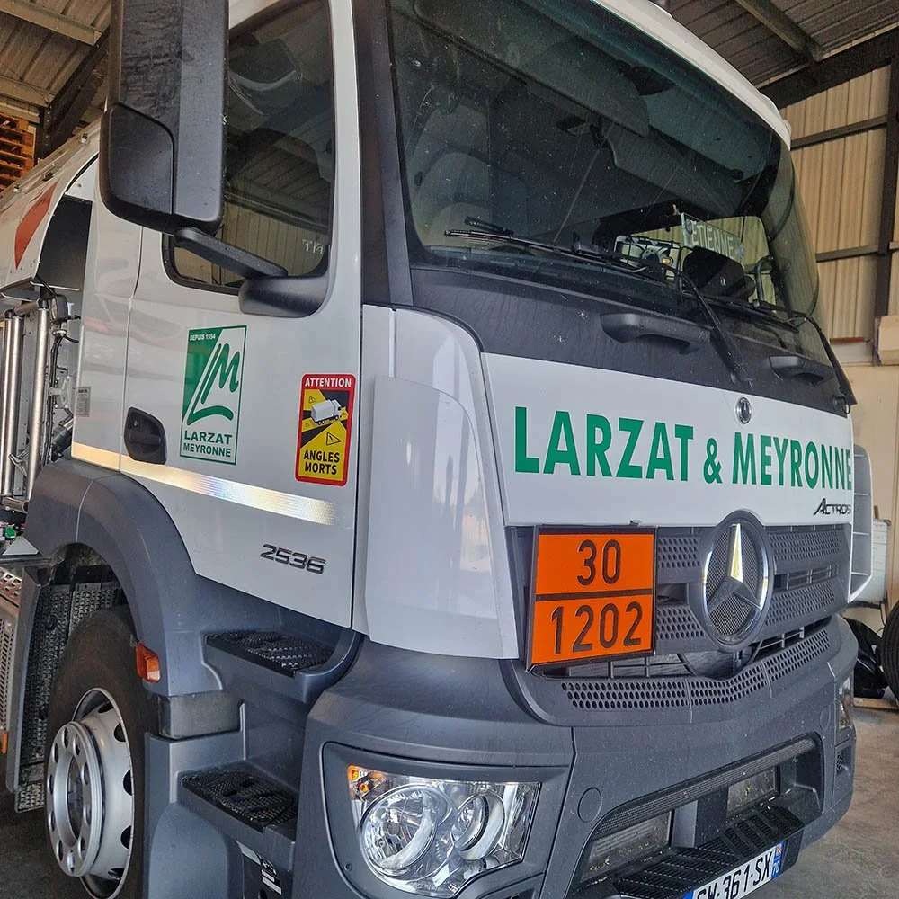 Un camion de service avec des étiquettes de danger orange et des inscriptions en vert et blanc "LARZAT & MEYRONNE" à l'avant, stationné à l'intérieur d'un hangar
