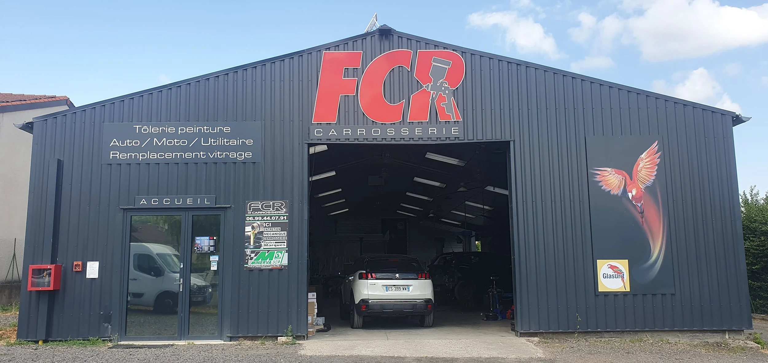 Façade d'un garage de carrosserie avec un grand panneau indiquant les services offerts, un affichage avec le numéro de contact et un logo de marque. Il y a une voiture blanche stationnée à l'intérieur, une fresque d'oiseau coloré sur la droite, et un