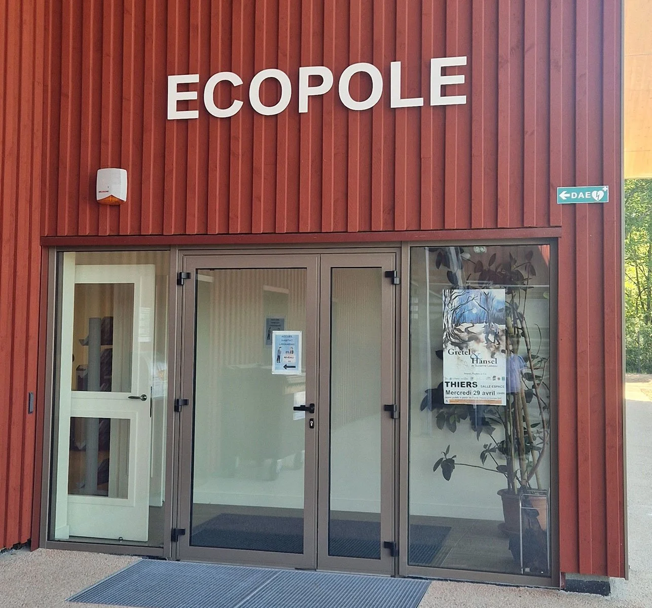 Entrée d'un bâtiment en bois avec le signe 'Ecopole' au-dessus, portes en verre, et une affiche promotionnelle à droite dans la fenêtre.