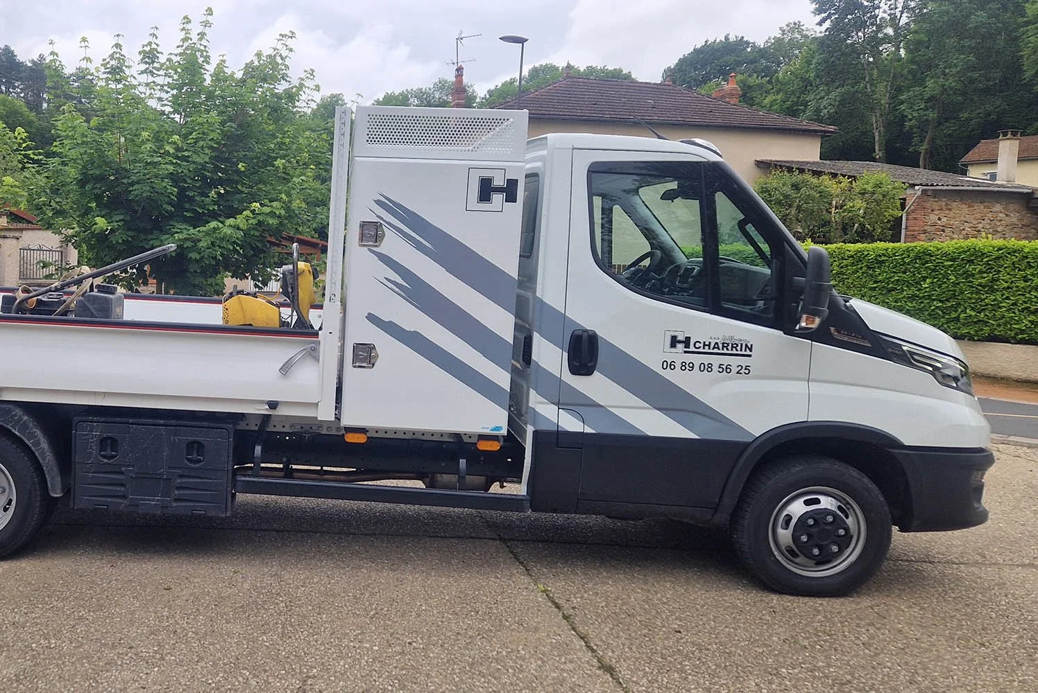 Un camion blanc de service avec un compartiment de rangement, stationné sur une route résidentielle bordée d'arbres et de maisons. Le camion porte le nom et le numéro de téléphone de l'entreprise "Charrin."