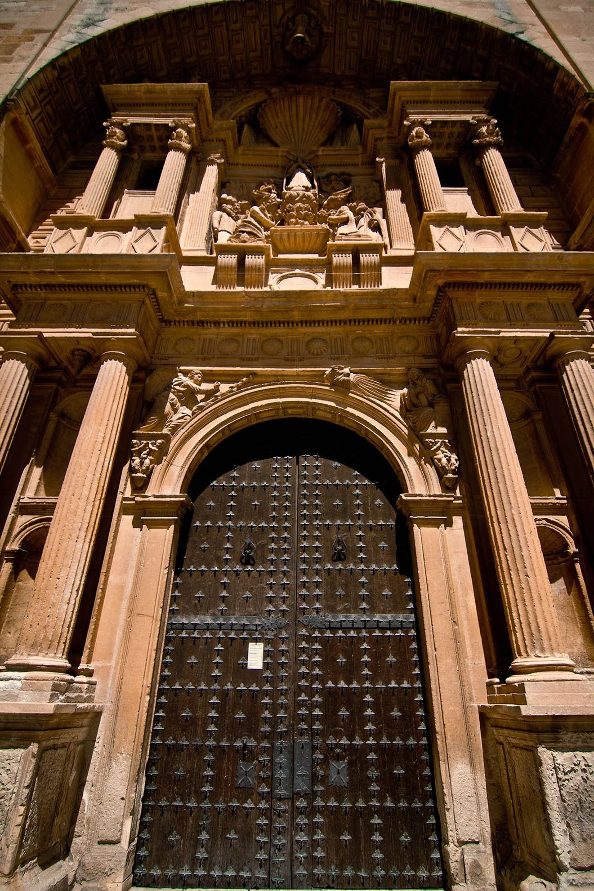 Vue d'une grande porte en bois ornée de clous de fer, entourée de colonnes en pierre et de sculptures détaillées, partie d'une façade architecturale ancienne.