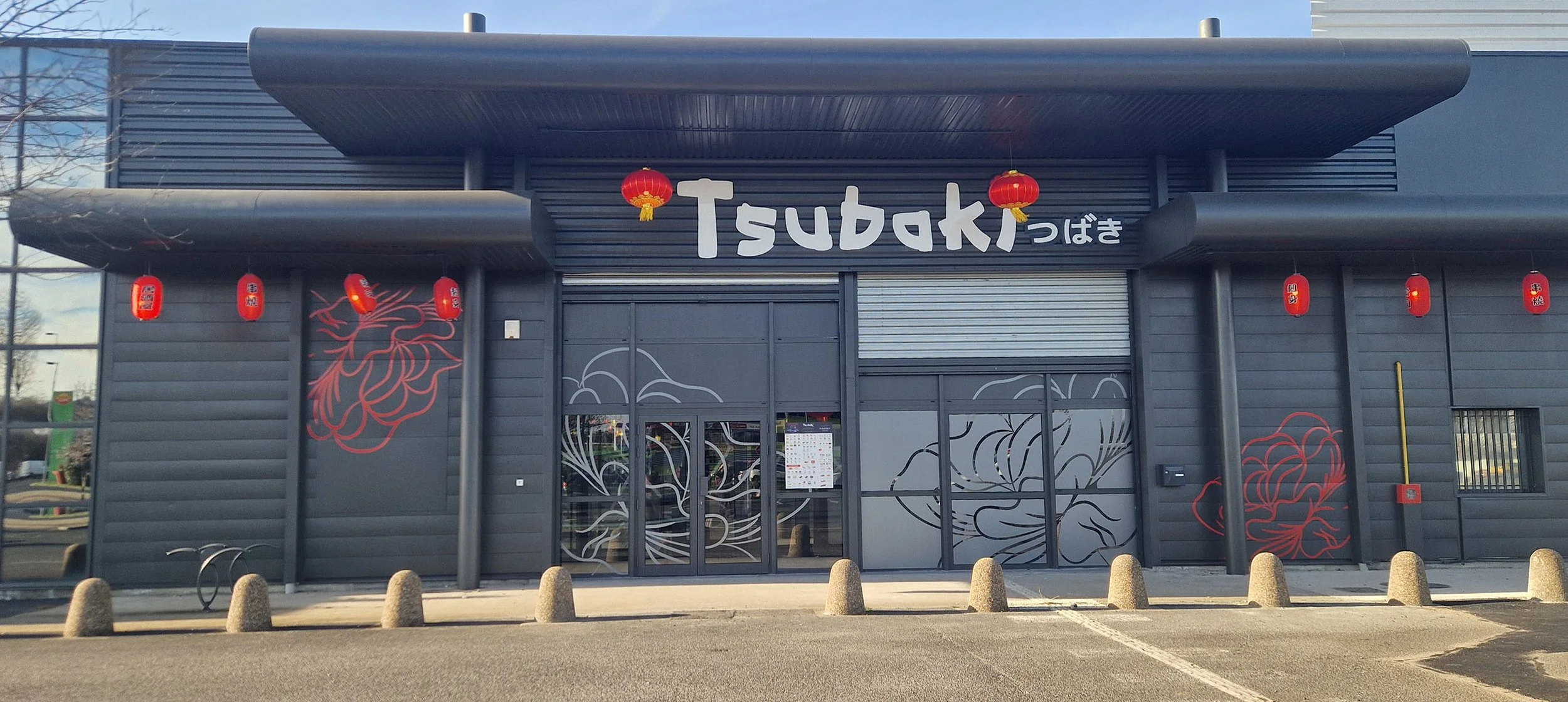 Façade d'un restaurant nommé Tsubaki, décorée de lanternes rouges et de motifs floraux rouges et noirs, avec un seuil à barrières en béton devant.
