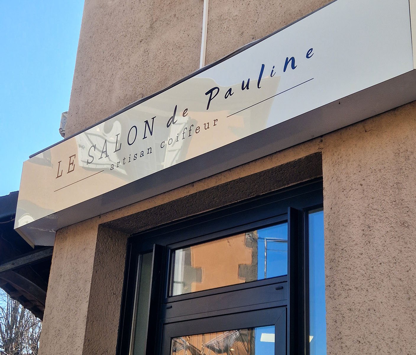 Panneau de salon de coiffure en français, indiquant "LE SALON de Pauline, artisan coiffeur" au-dessus d'une fenêtre à la façade beige.