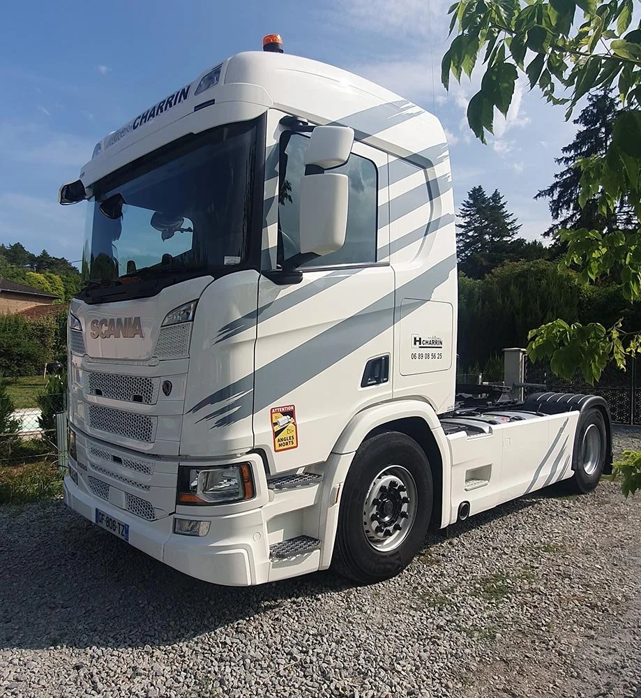 Un camion blanc de marque Scania, stationné sur un sol en gravier, avec des arbres et un ciel bleu en arrière-plan.