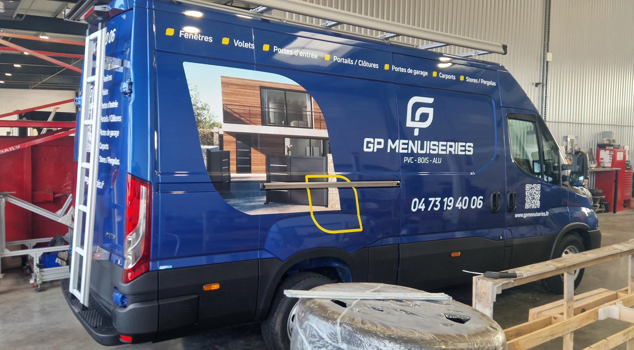 Camion de menuiserie bleu avec logo GPMenuiseries, affichant des images de travaux en bois, avec extensions et outils dans un atelier.