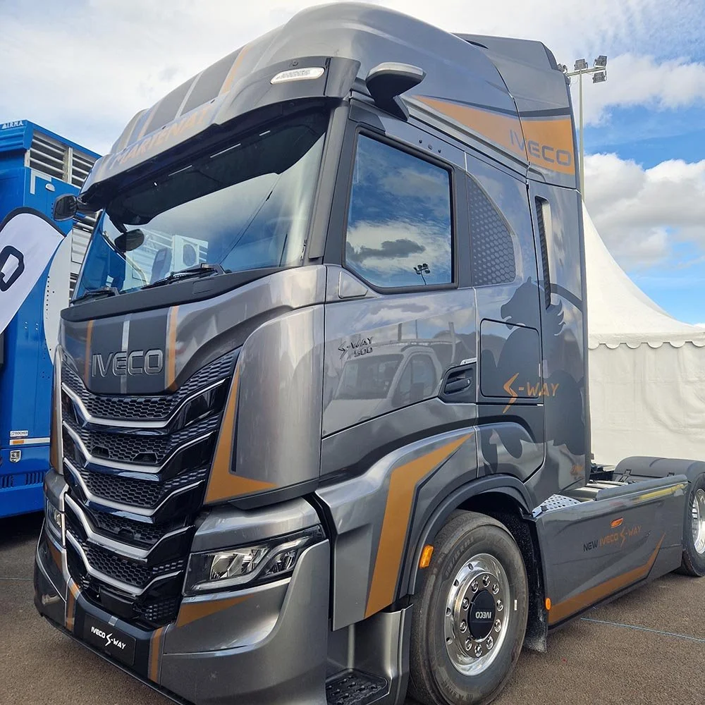 Un camion de couleur gris et orange, marque IVECO, avec des détails graphiques, parked devant un ciel partiellement nuageux lors d'un événement ou une exposition.