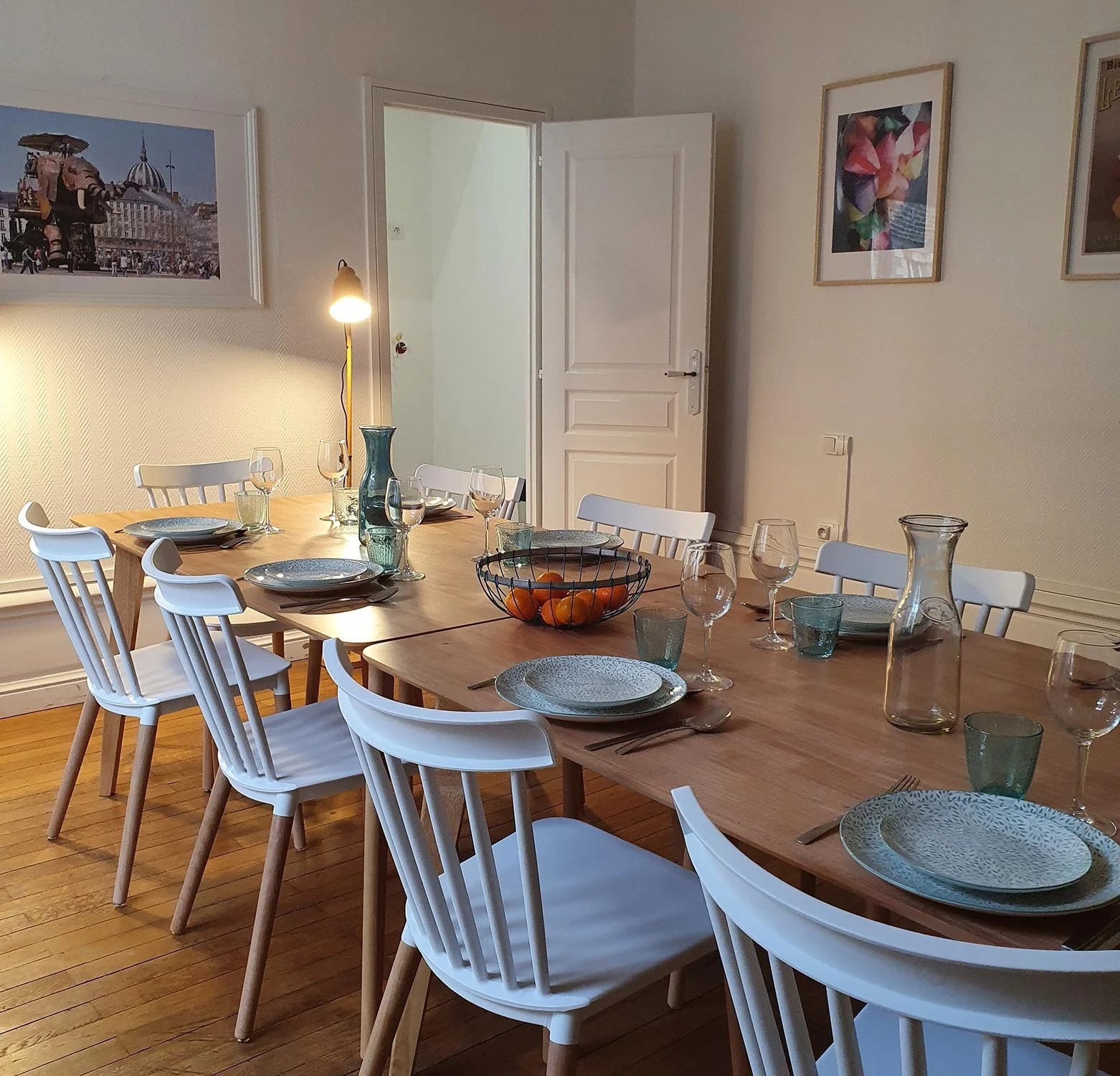 Salle à manger avec une grande table en bois, entourée de chaises blanches, dressée avec des assiettes, des verres, des couverts, et un bol de mandarines. La pièce est décorée de plusieurs tableaux et a un éclairage chaleureux.