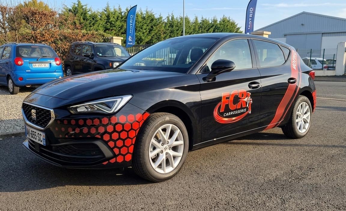 Voiture noire avec logo FCR Carrosseries sur la porte, stationnement en extérieur, autres véhicules en arrière-plan, bâtiments et drapeaux promotionnels.