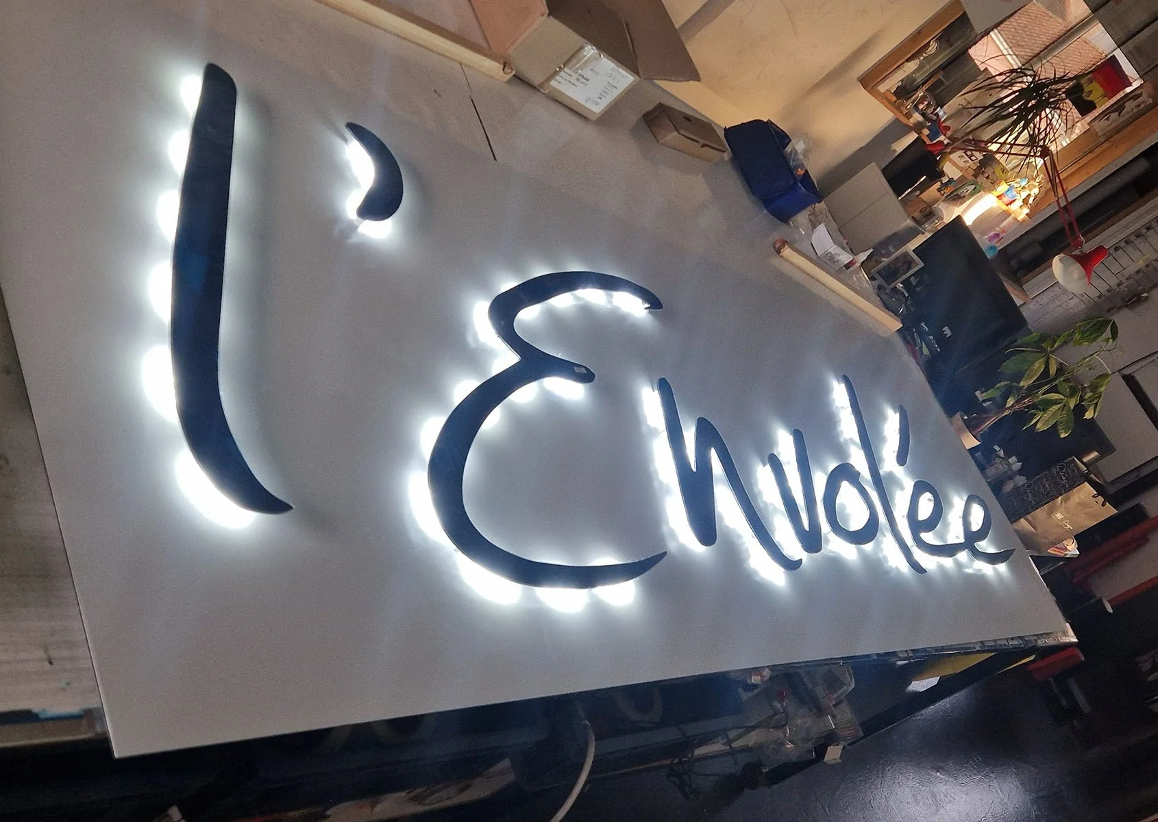 Un panneau lumineux avec le texte "Hello" écrit en script, entouré de lumières LED blanches, dans un environnement intérieur avec des meubles et des plantes.