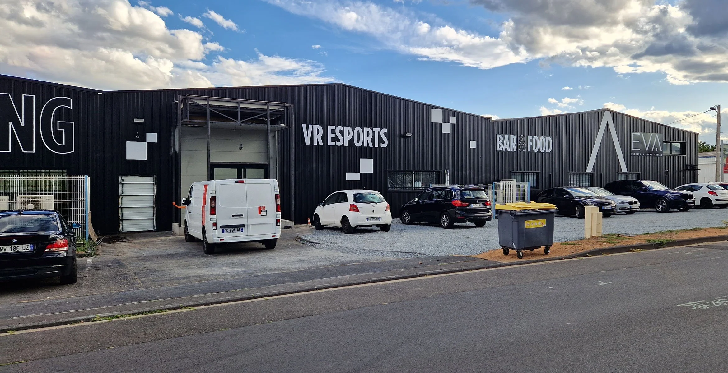 Bâtiment noir avec des inscriptions : VR ESPORTS, BAR & FOOD, EA. Parking avec plusieurs voitures dont une blanche, une noire, une gris clair et une noire, ainsi qu'une poubelle grise avec un couvercle jaune en dehors du bâtiment. Ciel partiellement 