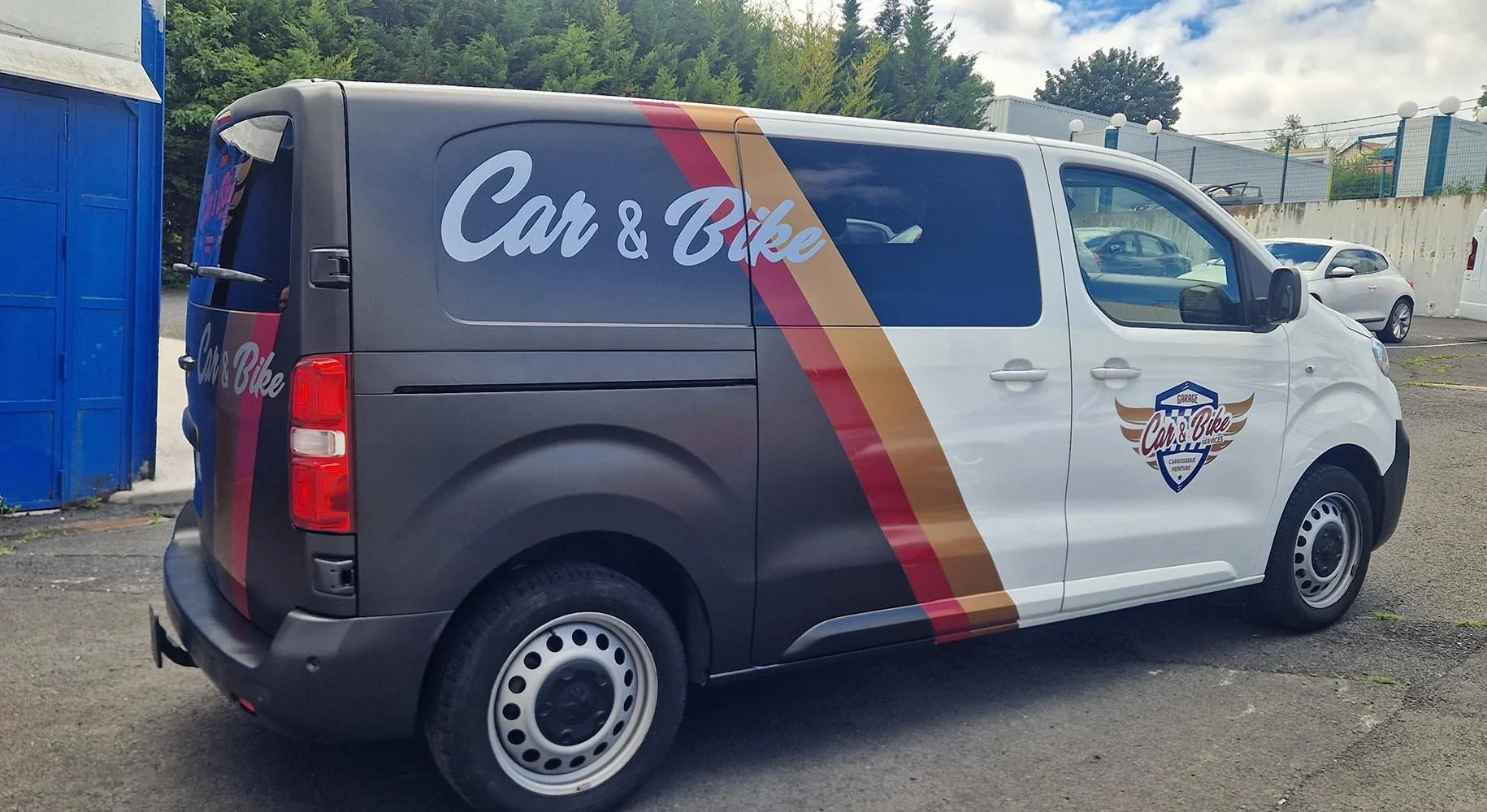 Véhicule de service pour Car & Bike, garé dans un parking avec d'autres voitures, en arrière-plan un mur en béton et des arbres.