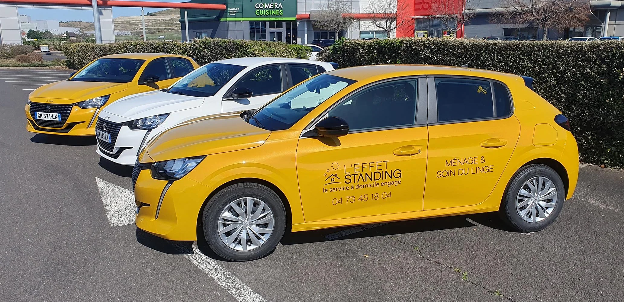 Voiture jaune avec publicité pour un service de ménage à domicile, stationnée dans une zone de parking, avec deux autres voitures blanches et jaunes à côté, devant un centre commercial.