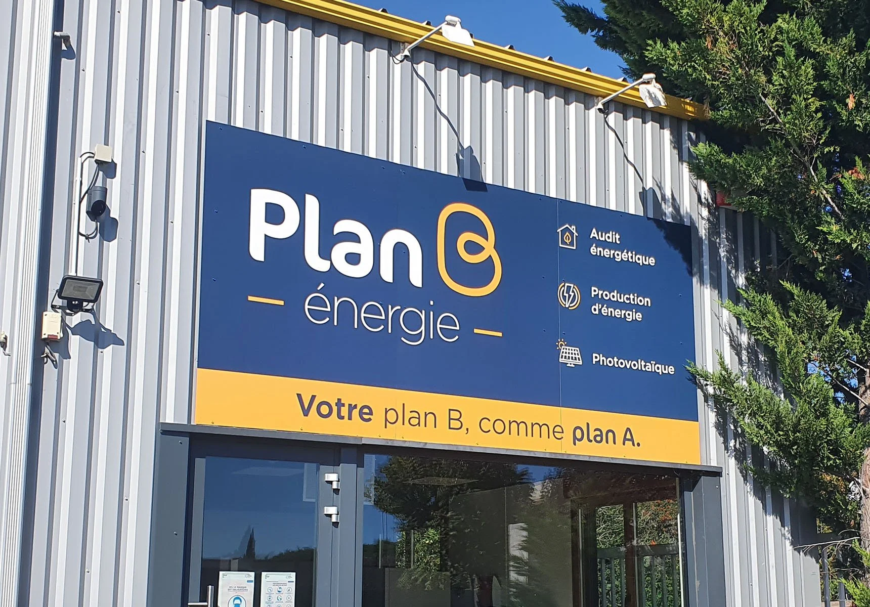 Une façade d'immeuble avec un panneau publicitaire bleu et jaune indiquant 'Plan énergie' avec des icônes pour audit énergétique, production d'énergie et photovoltaïque, ainsi que la phrase 'Votre plan B, comme plan A'.