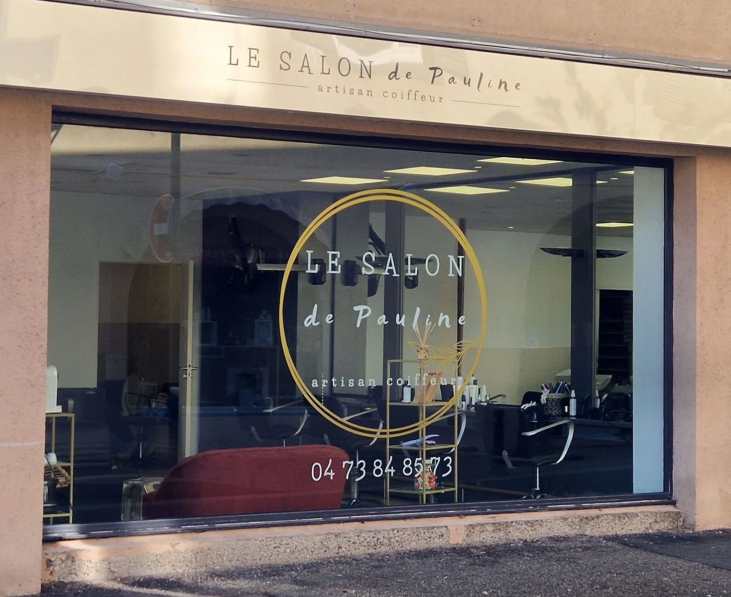 Vitrine d'un salon de coiffure nommé 'Le Salon de Pauline'. Description du lieu visible à travers la vitre, avec des fauteuils de coiffure et des produits capillaires. Le numéro de téléphone est inscrit en bas.