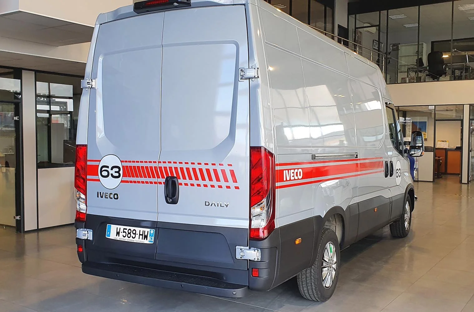 Un camion blanc Iveco Daily avec des marques rouges, stationné à l'intérieur d'un bâtiment, avec un logo d'entreprise et un numéro 63 sur la coque.