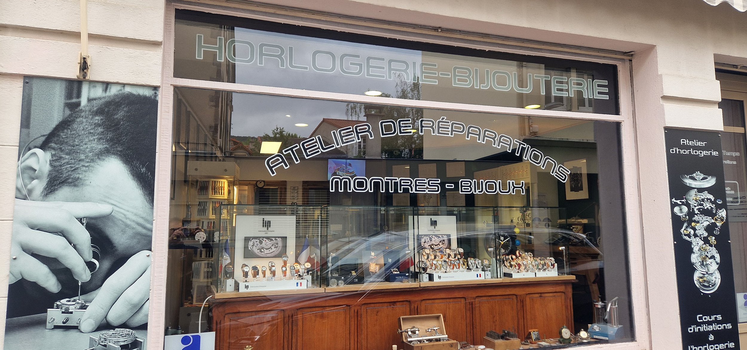 Vitrine d'un atelier de réparation horlogerie-bijouterie avec affiches de montres et de mécanismes d'horlogerie, et une grande photo en noir et blanc d'une personne manipulant une montre.