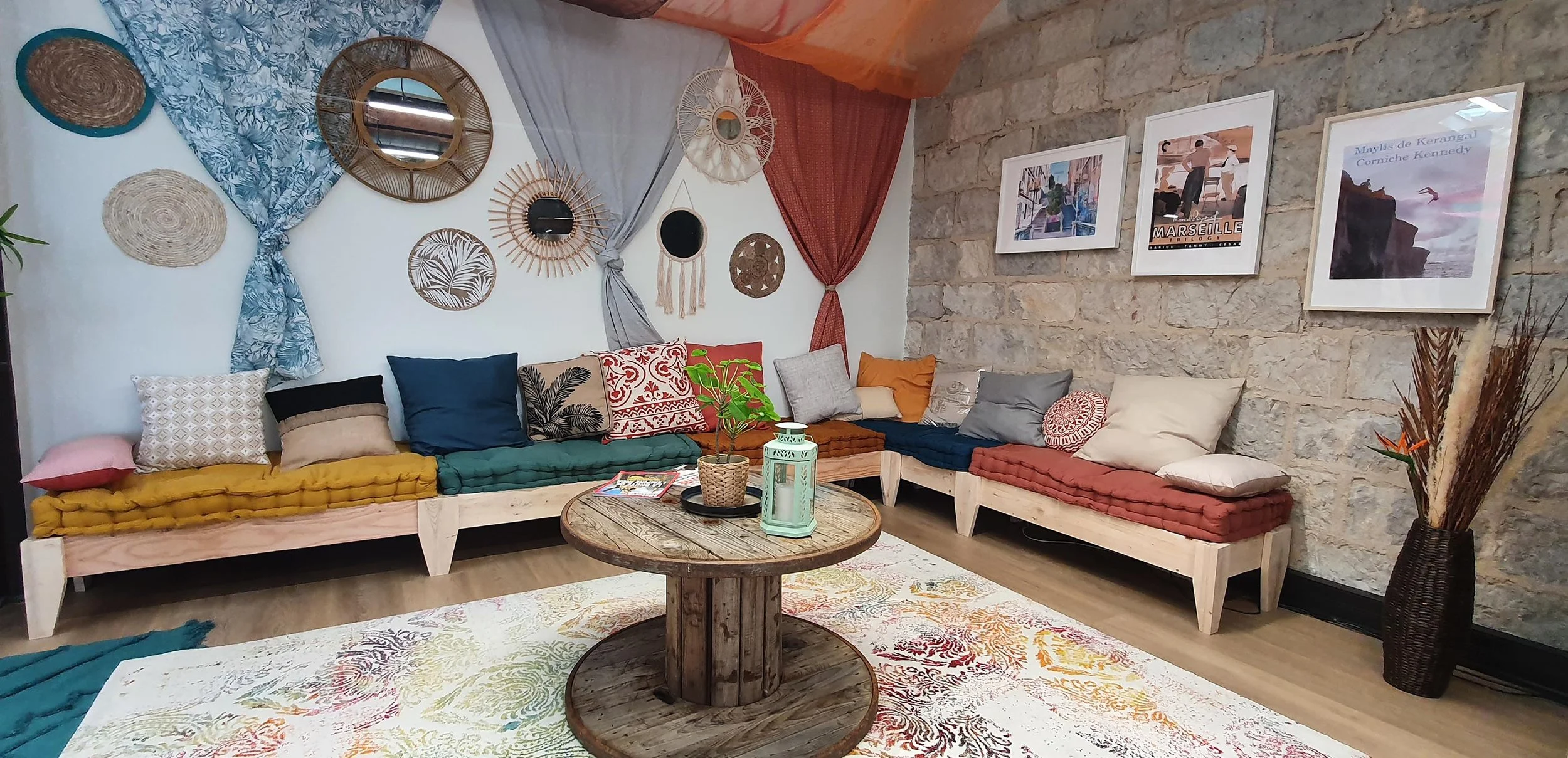 Salon de détente avec canapés en bois avec coussins colorés, table ronde en bois, mur en pierre avec photos encadrées, décoration bohème avec miroirs, paniers, et plantes.